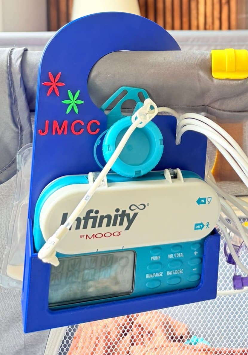 The holder hanging on a crib rail by its arch handle, with the pump running and the feeding line in place.